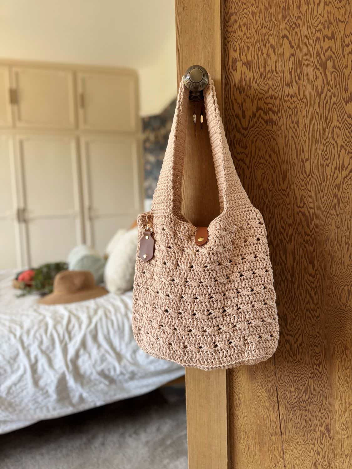 A beige crochet cross body bag, crafted from a free pattern and adorned with a leather tag, hangs on a wooden door; a bed and wardrobe are visible in the softly lit background.