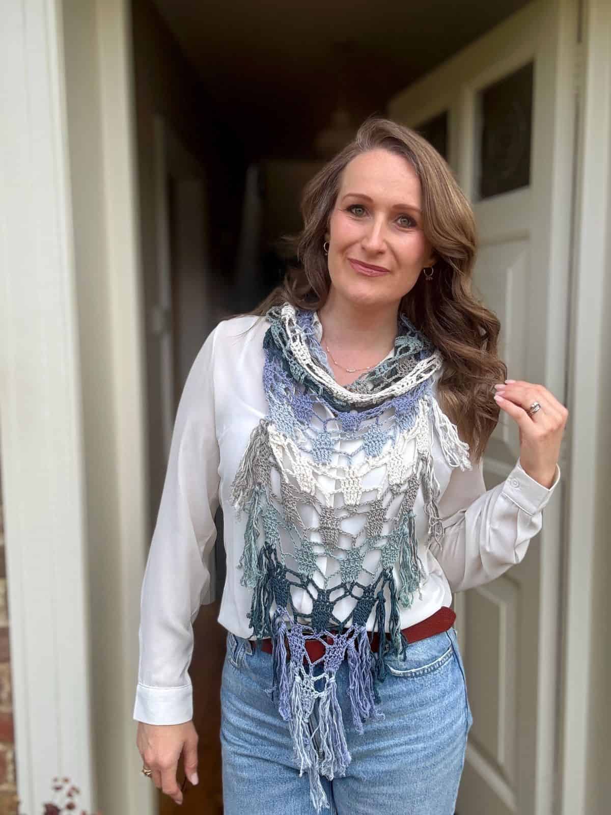 Woman standing in a doorway, wearing a white blouse, blue jeans, and a multicolored bandana scarf crochet pattern with fringe detail.