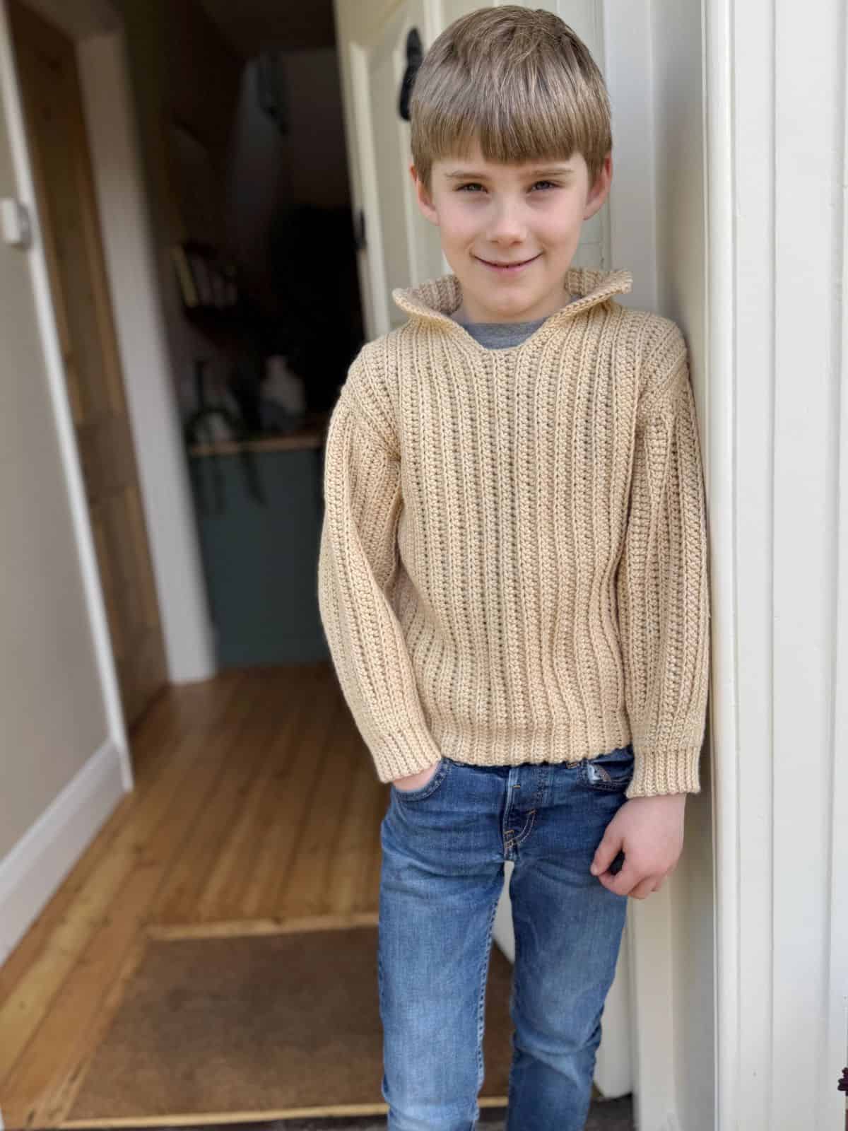 A young boy with short light brown hair, wearing a beige crochet sweater made from easy crochet sweater patterns for beginners and blue jeans, stands indoors near a doorway on a wooden floor.