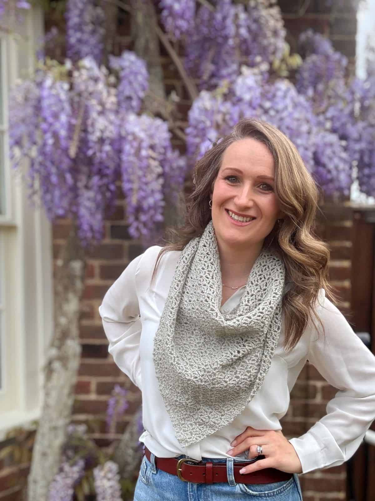 Woman standing outdoors in front of a brick wall and blooming wisteria, wearing a white blouse, jeans, and a delicate lace crochet shawl pattern as a scarf, smiling at the camera.