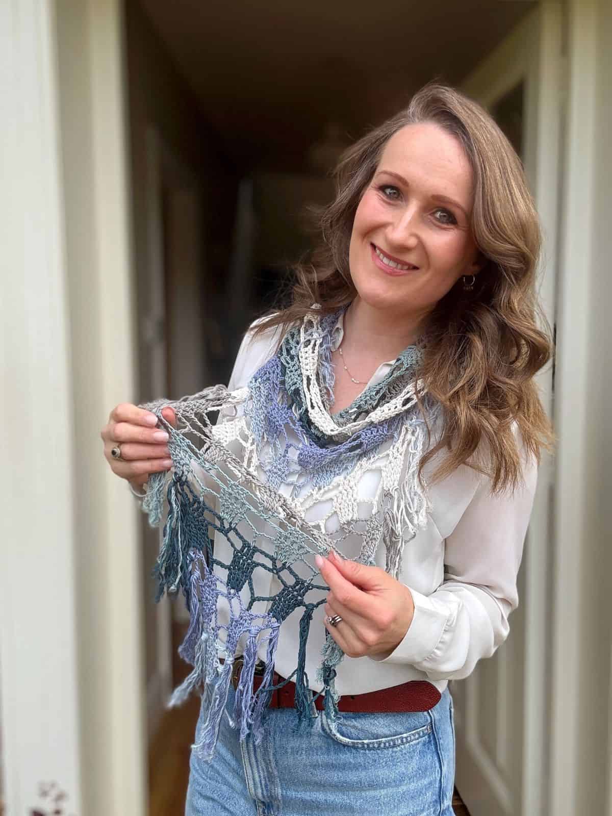 Woman with wavy hair wearing a white blouse and blue jeans stands indoors, smiling and holding a handmade bandana scarf crochet pattern in blue, grey, and white tones.
