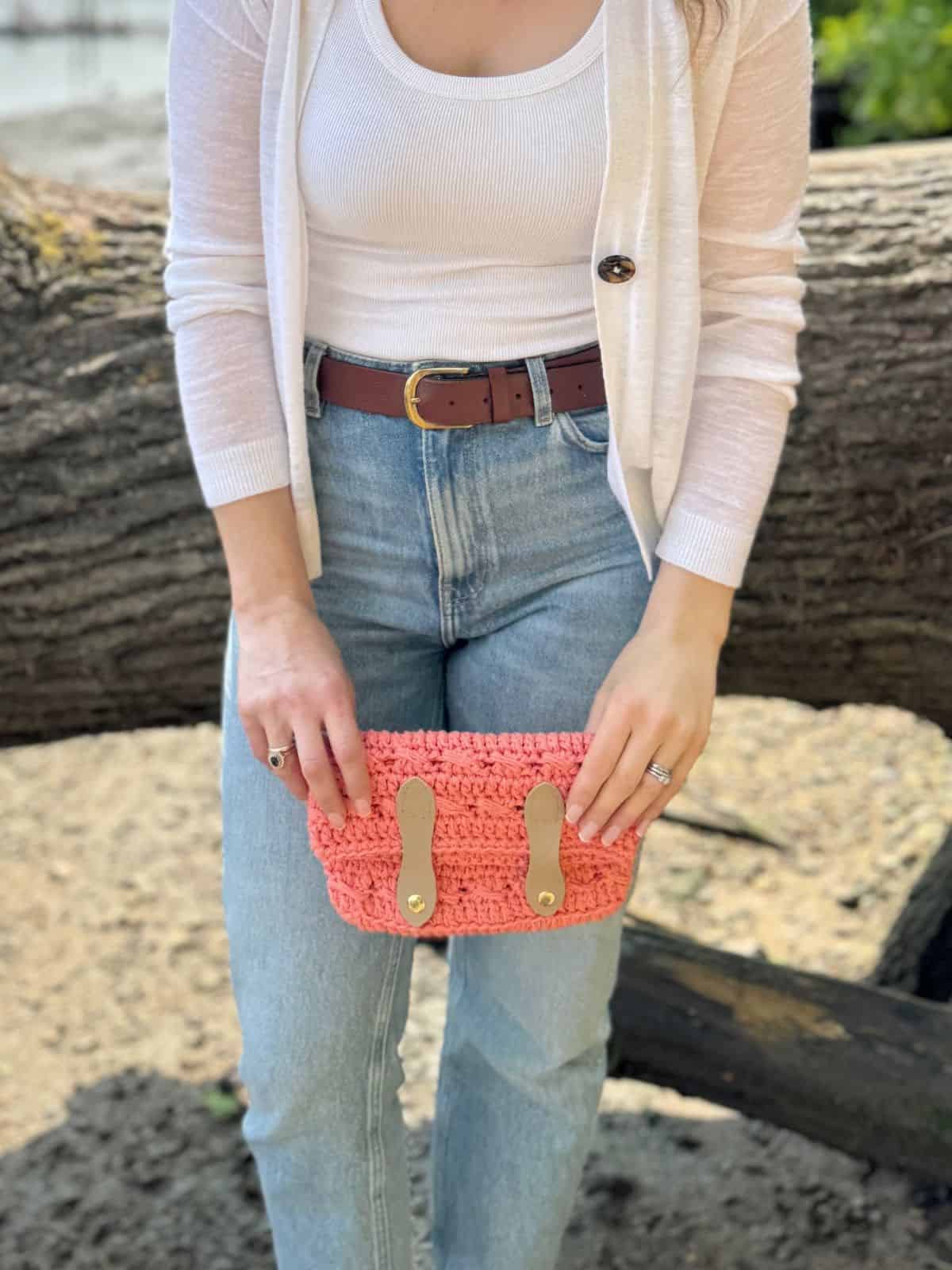 Woman in a white top and light cardigan holds a pink crochet clutch bag, crafted using a stylish crochet clutch bag pattern, with leather straps. She stands outdoors in front of a fallen tree.