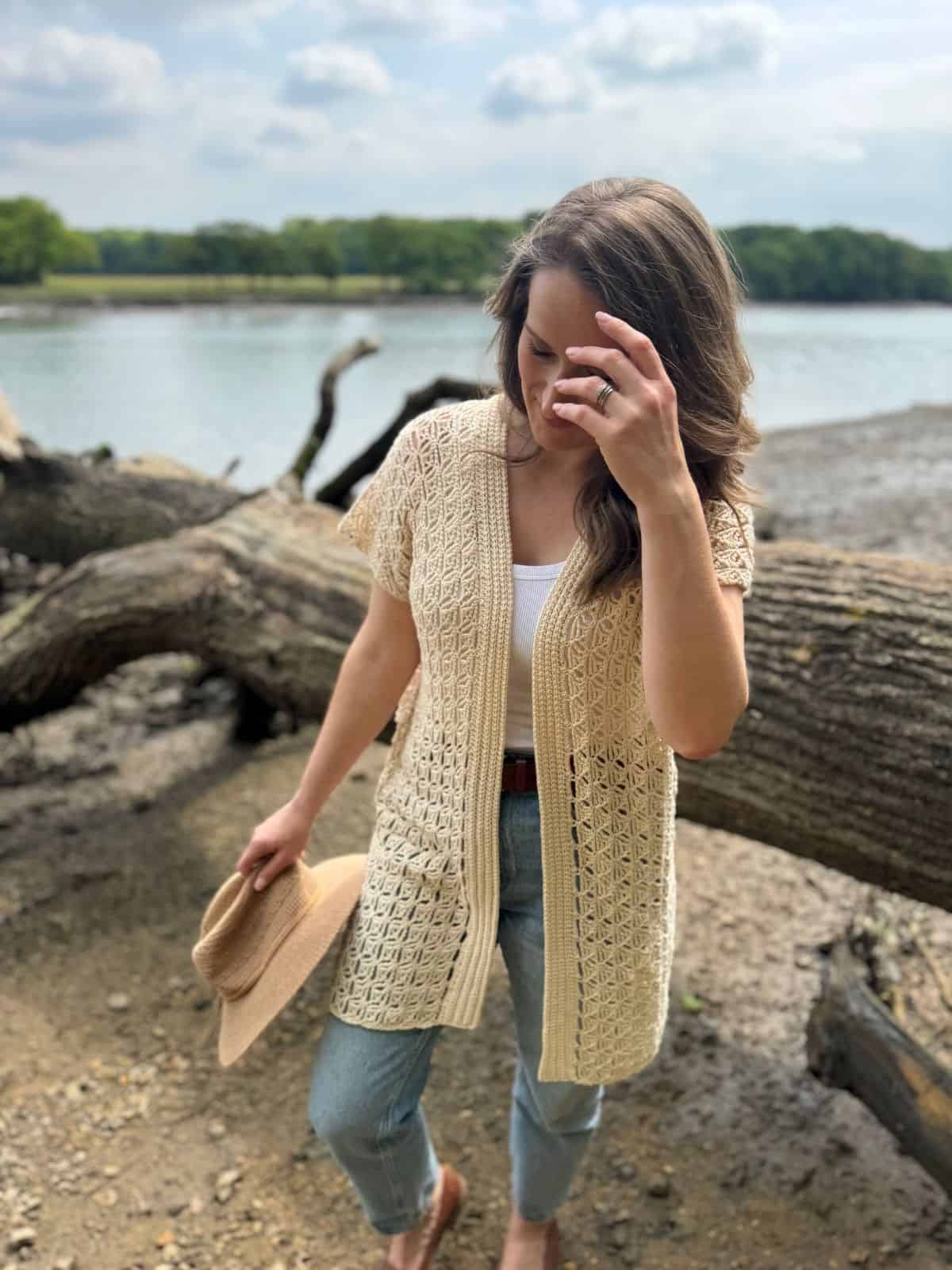 A woman stands by a lake, holding a hat and touching her hair. She is wearing a cream summer cardigan crochet pattern over a white top and blue jeans. Fallen tree limbs are in the background.