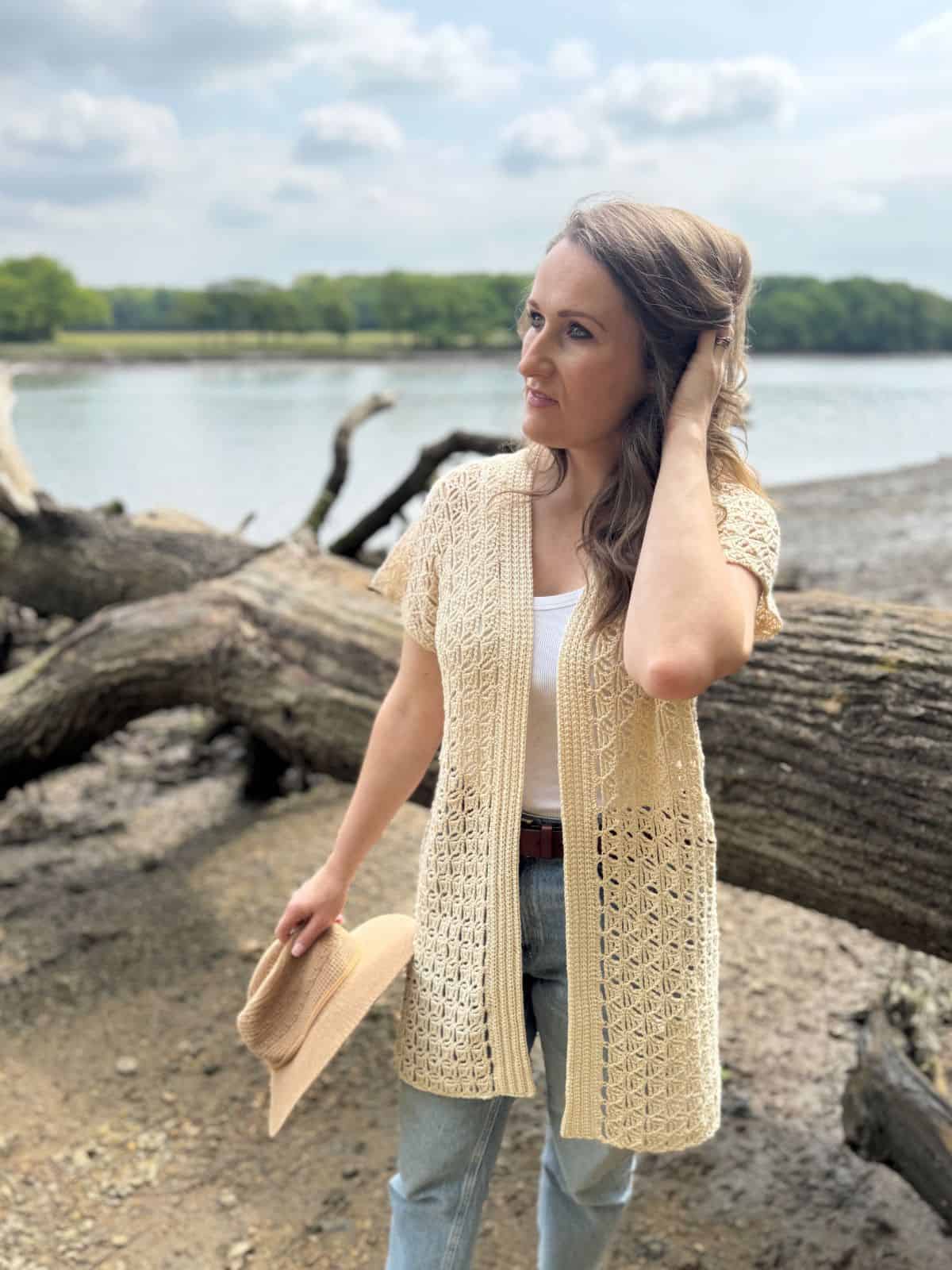 A woman stands outdoors by a lakeshore, holding a hat in one hand and touching her hair with the other. She wears a light summer cardigan crochet pattern over a white top and jeans.