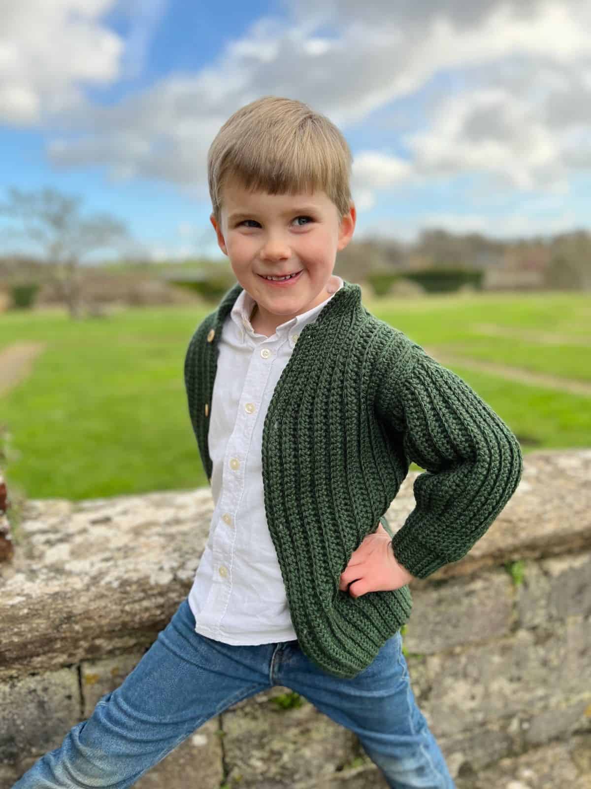 A young boy in a white shirt, green crocheted cardigan made with easy crochet sweater patterns for beginners, and jeans stands outdoors by a stone wall with green grass and a cloudy sky in the background.