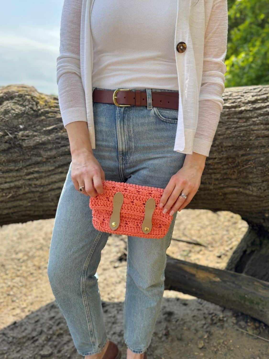 Person wearing light jeans, a white top, and a cardigan stands outdoors holding a coral crochet cross body bag—find the free pattern online. A large tree trunk is in the background.