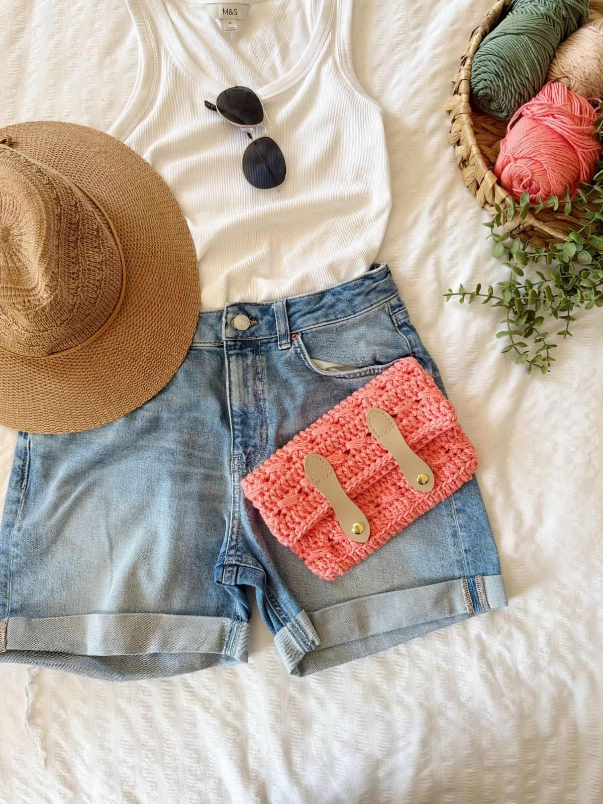 A summer outfit with a straw hat, white tank top, sunglasses, denim shorts, a pink crochet clutch bag pattern, and a basket of yarn on a white textured surface.