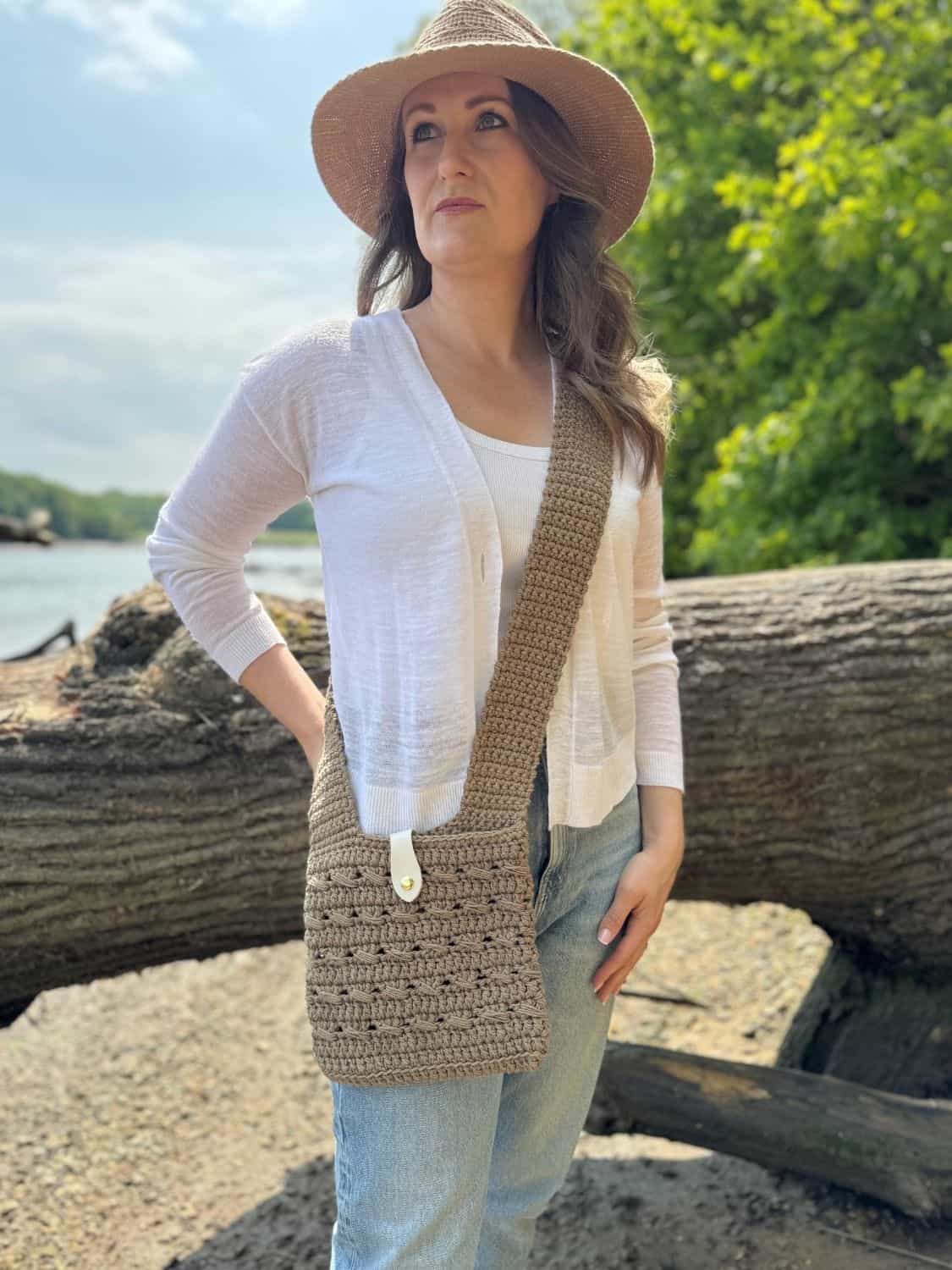 Woman standing outdoors by a large fallen tree, wearing a straw hat, white cardigan, jeans, and carrying a crochet crossbody bag made from a free pattern.