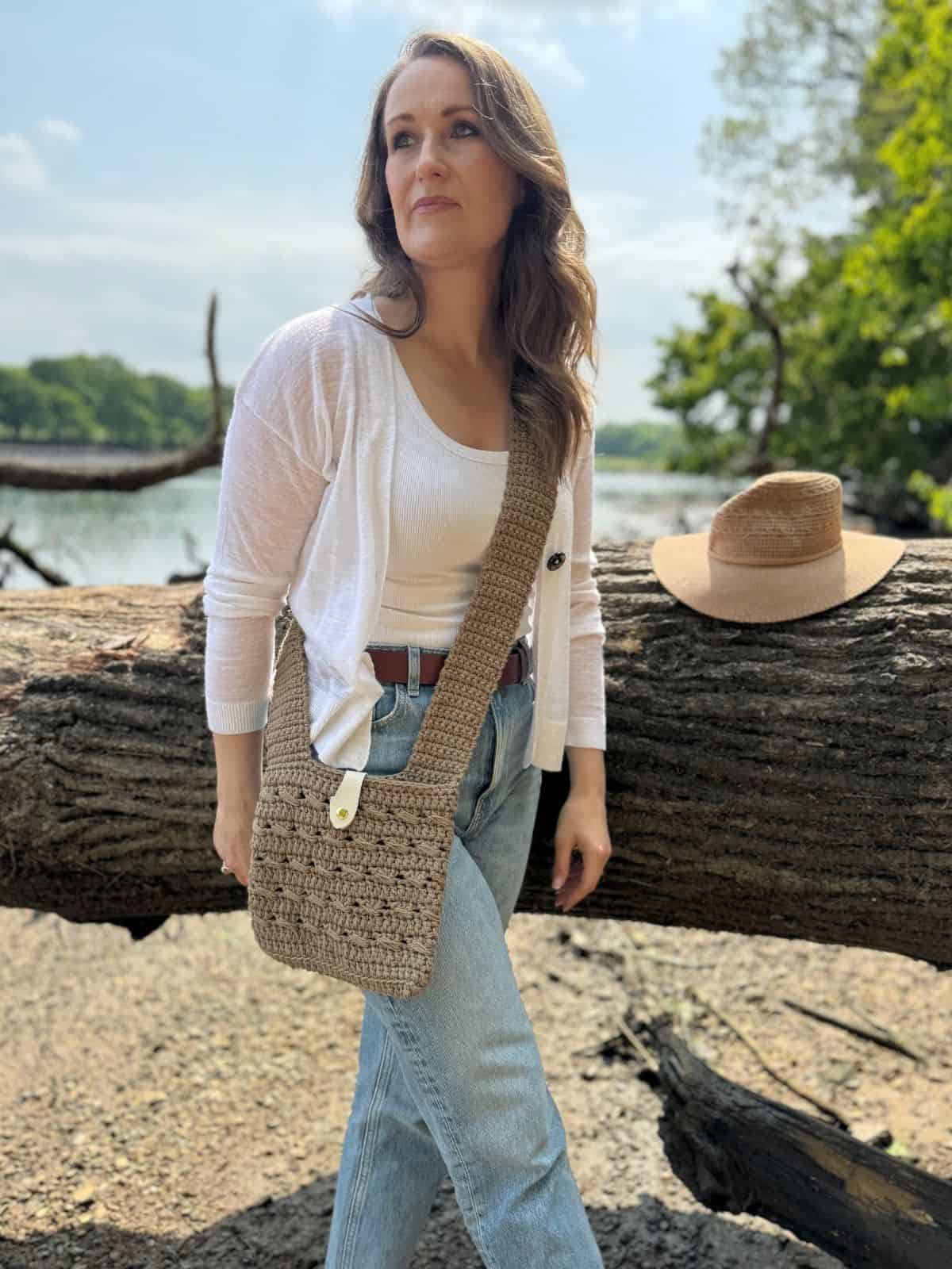 A woman stands outdoors by a fallen tree, wearing a white top, light blue jeans, and a crochet cross body bag. A straw hat rests on the tree beside her. Trees and water are in the background—perfect inspiration for a crochet cross body bag free pattern.