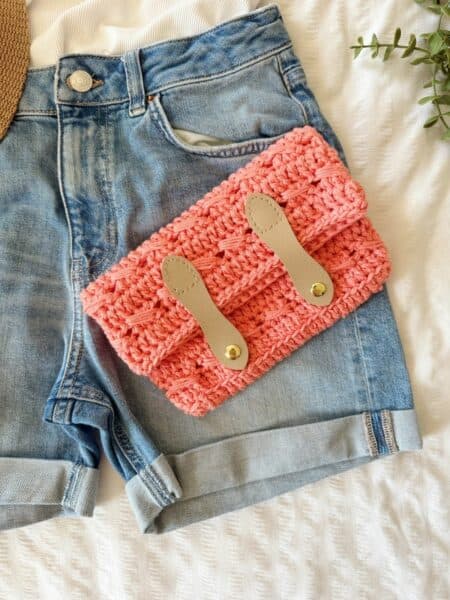 A pair of denim shorts with a crocheted pink clutch, crafted using a crochet clutch bag pattern and featuring tan leather straps, sits on a white textured surface.