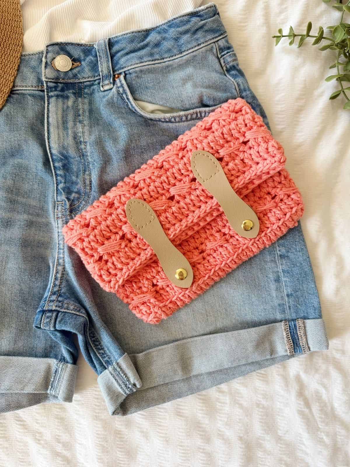 A pair of denim shorts with a crocheted pink clutch, crafted using a crochet clutch bag pattern and featuring tan leather straps, sits on a white textured surface.