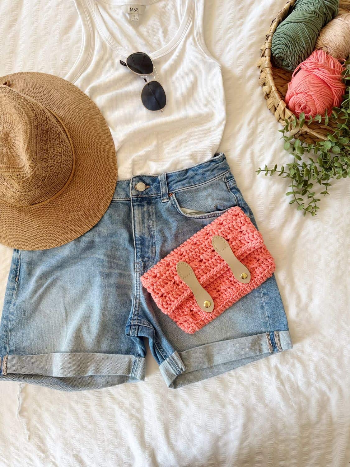Flat lay of a summer outfit with a straw hat, sunglasses, white tank top, denim shorts, and a pink crochet cross body bag free pattern clutch next to a basket of yarn on a white textured surface.