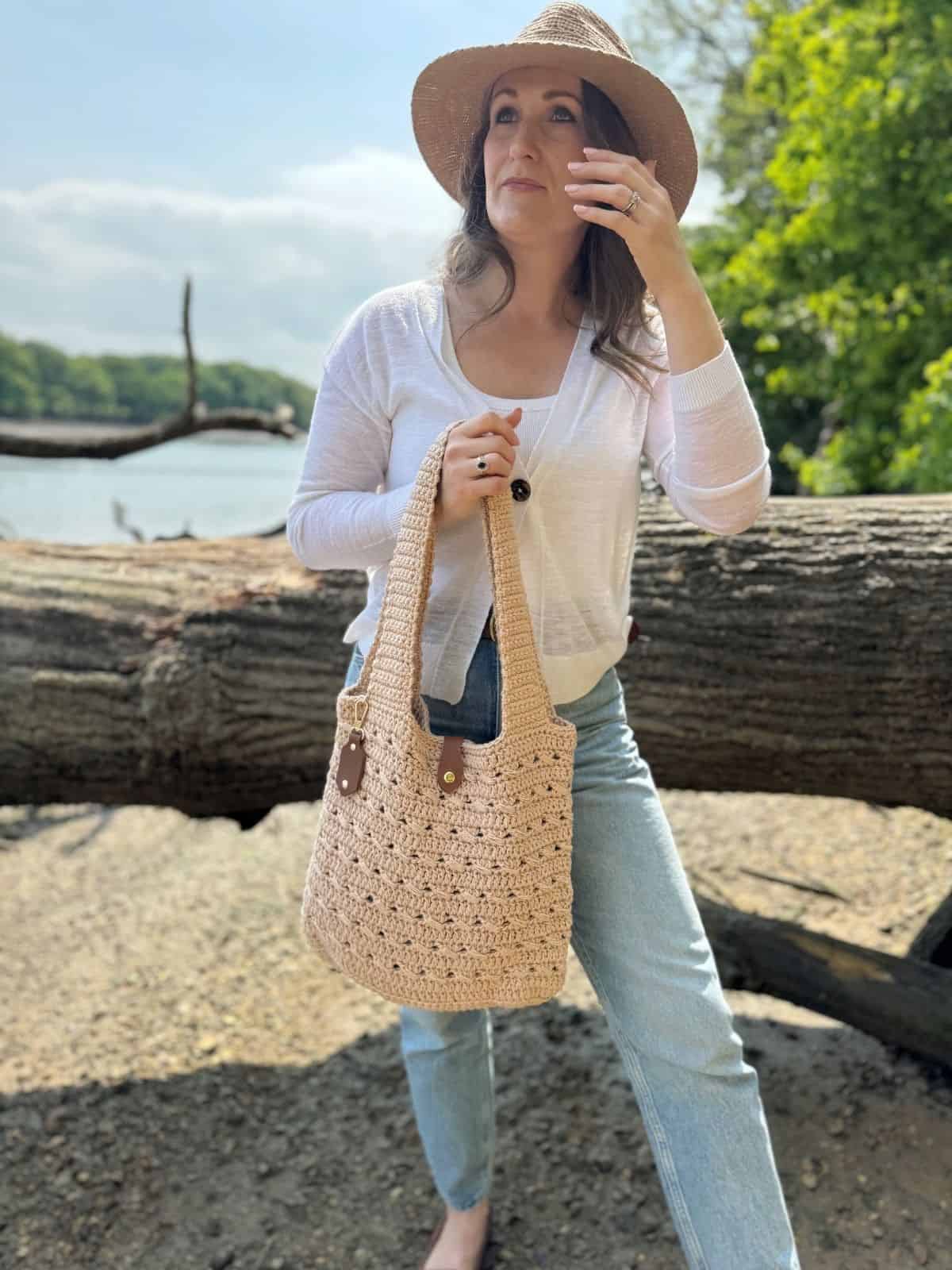 Woman wearing a straw hat, white top, and jeans stands outdoors by a fallen tree, holding a tote made from a free crochet market bag pattern—with greenery and water in the background.