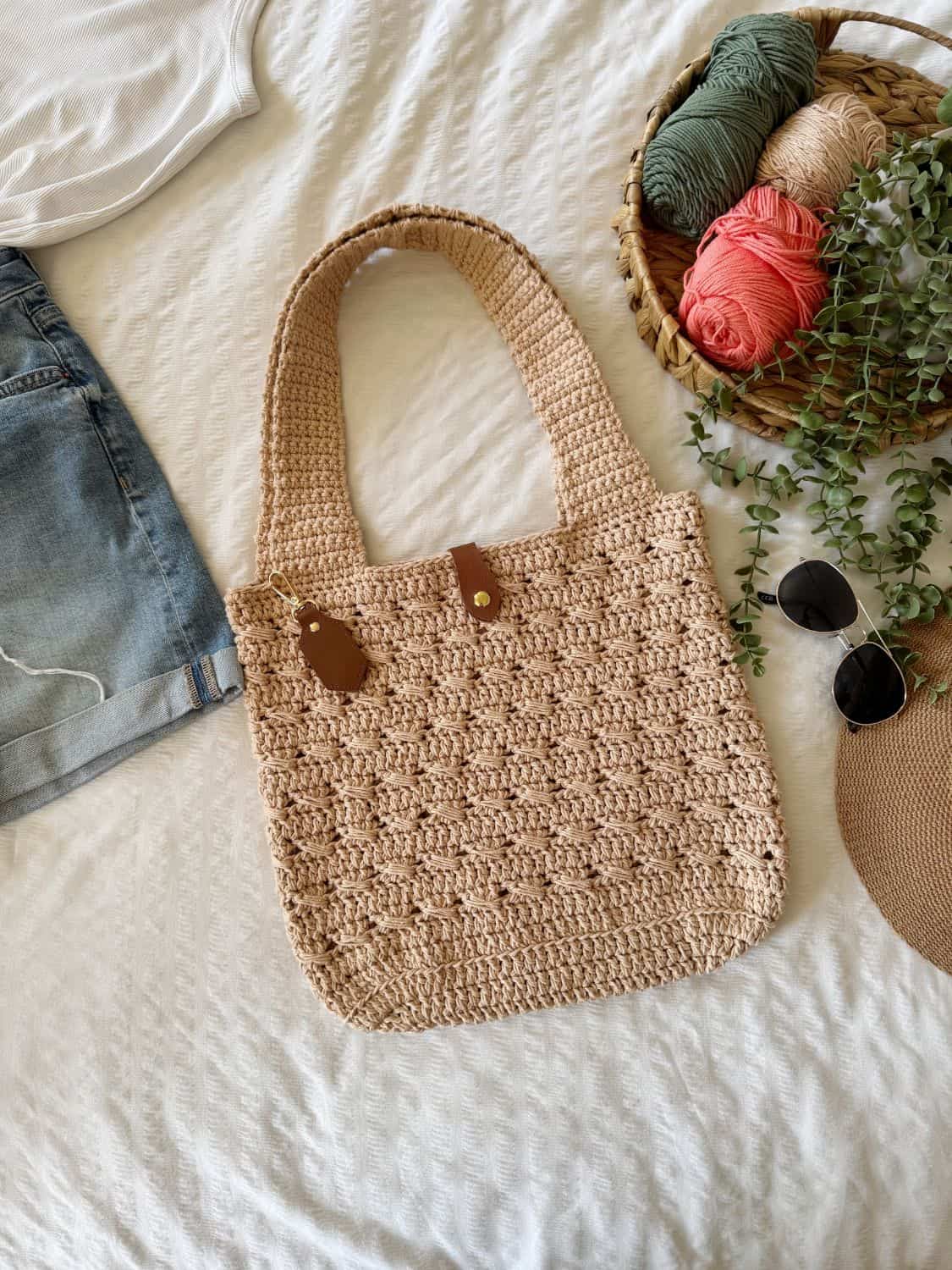 A beige crocheted tote bag, inspired by a crochet cross body bag free pattern, is laid on a bed next to denim shorts, a white shirt, sunglasses, a hat, and a basket of yarn and greenery.