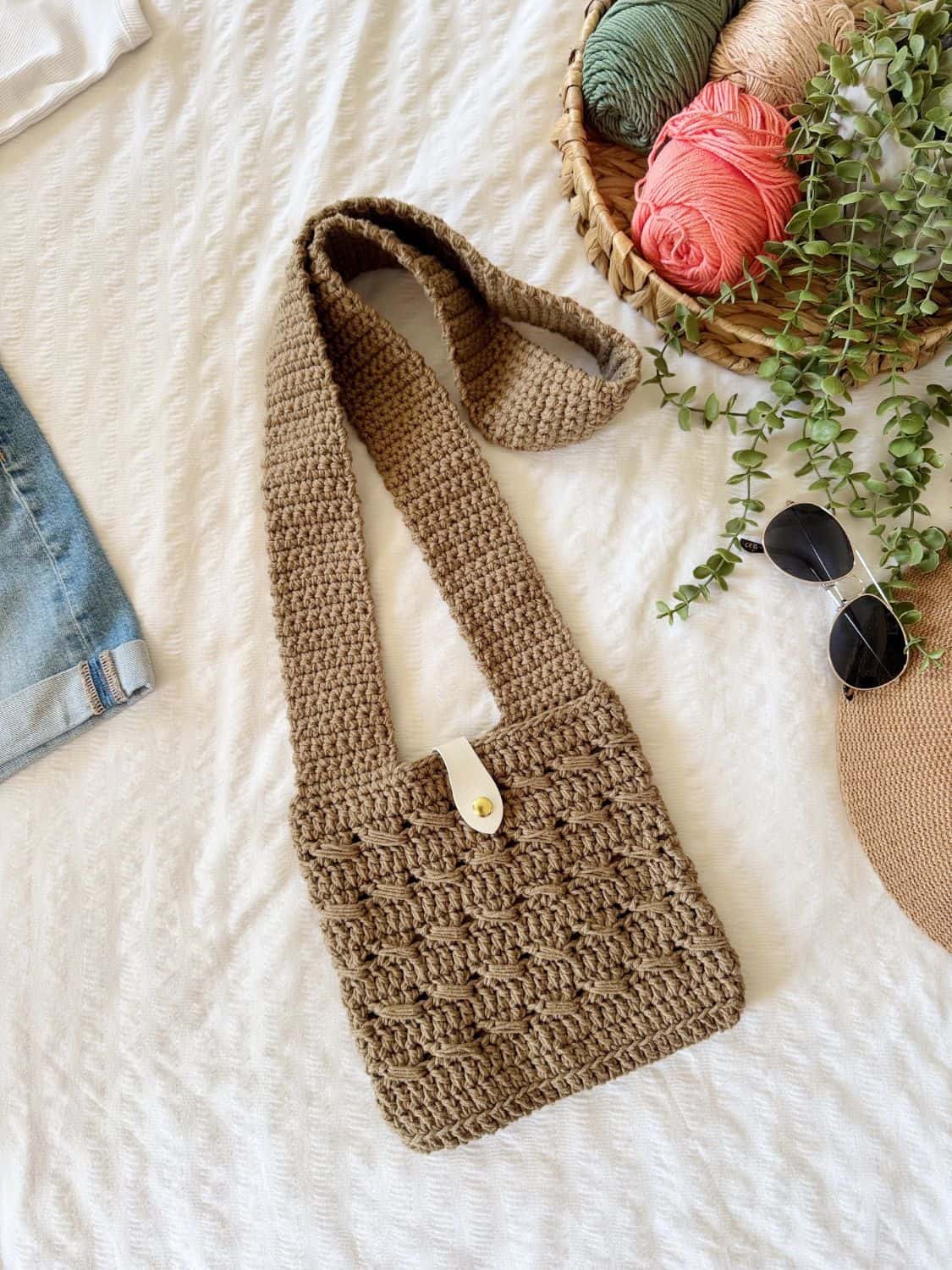 A brown crochet cross body bag, featuring a button closure, rests on a white textured surface beside yarn, greenery, sunglasses, and a woven hat—perfect inspiration for your next project or to find a crochet cross body bag free pattern.