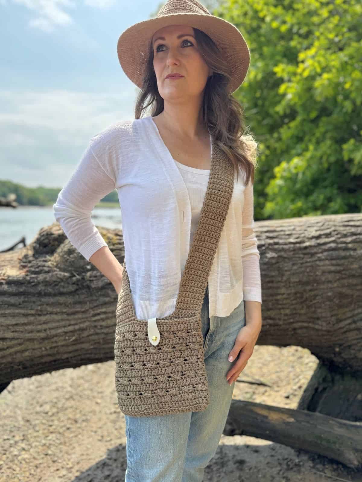 Woman standing outdoors by a fallen tree, wearing a straw hat, white top, blue jeans, and carrying a stylish crochet cross body bag—check out our free pattern to make your own.