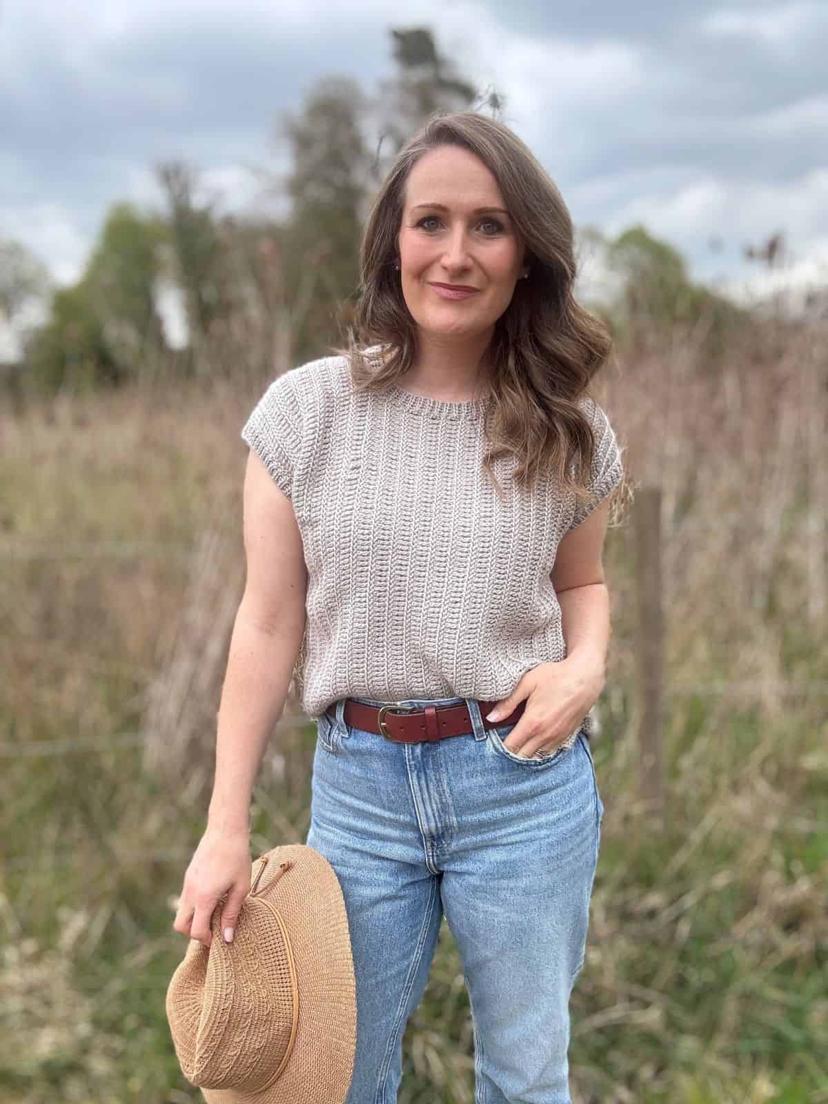 A woman stands outdoors in a field, wearing a light crochet summer top pattern and blue jeans, holding a straw hat in one hand, with trees and a cloudy sky in the background.