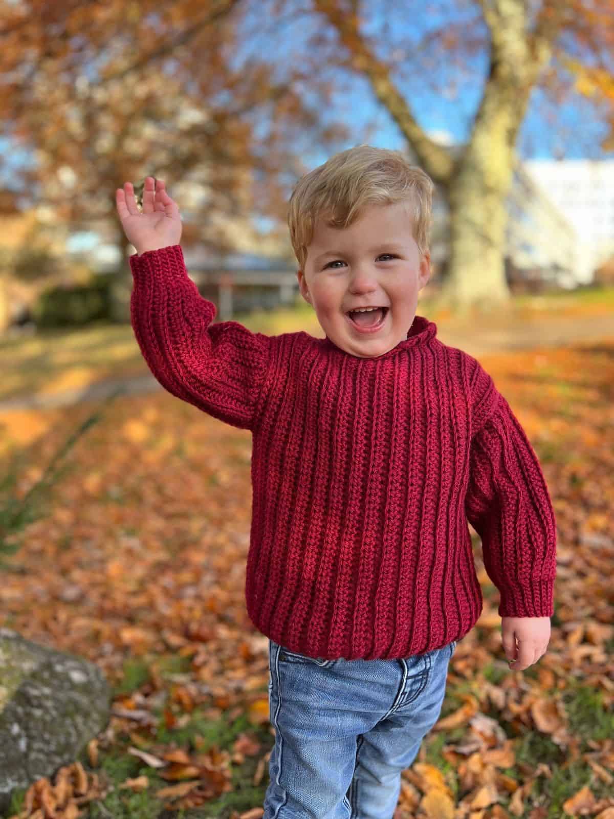Young child in a red crochet sweater, crafted from easy crochet sweater patterns for beginners, stands outdoors on autumn leaves, smiling and waving with one hand raised.