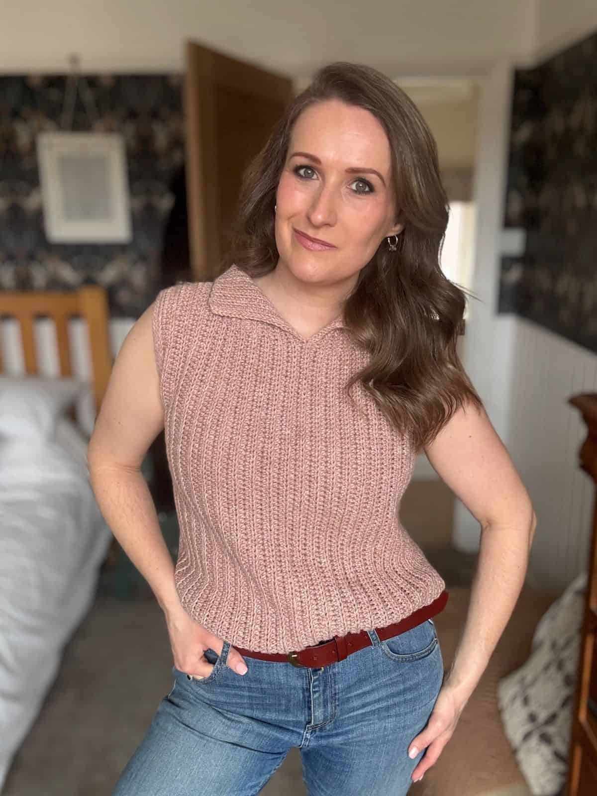 A woman with long brown hair wearing a modern crochet vest and jeans stands in a bedroom, looking at the camera with a neutral expression.