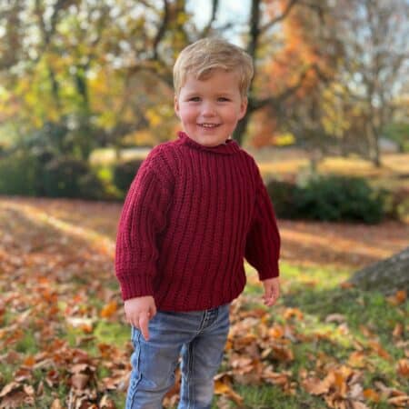 A young child stands outdoors on grass covered with autumn leaves, wearing a red crochet sweater, jeans, and brown shoes, with trees and fall foliage in the background.