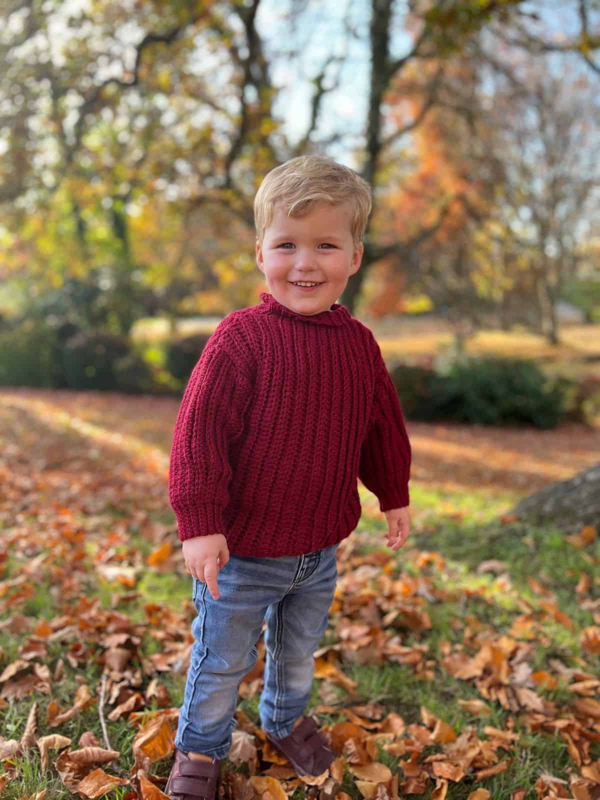 A young child stands outdoors on grass covered with autumn leaves, wearing a red crochet sweater, jeans, and brown shoes, with trees and fall foliage in the background.