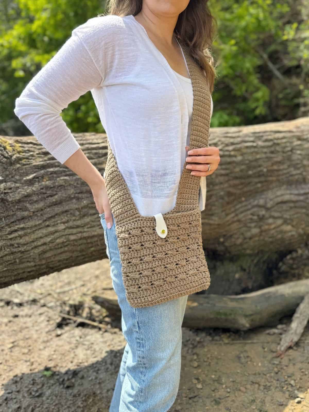 A person wearing a white cardigan and jeans stands outdoors with a beige crochet cross body bag, perfect for those seeking a free pattern to make their own stylish accessory.