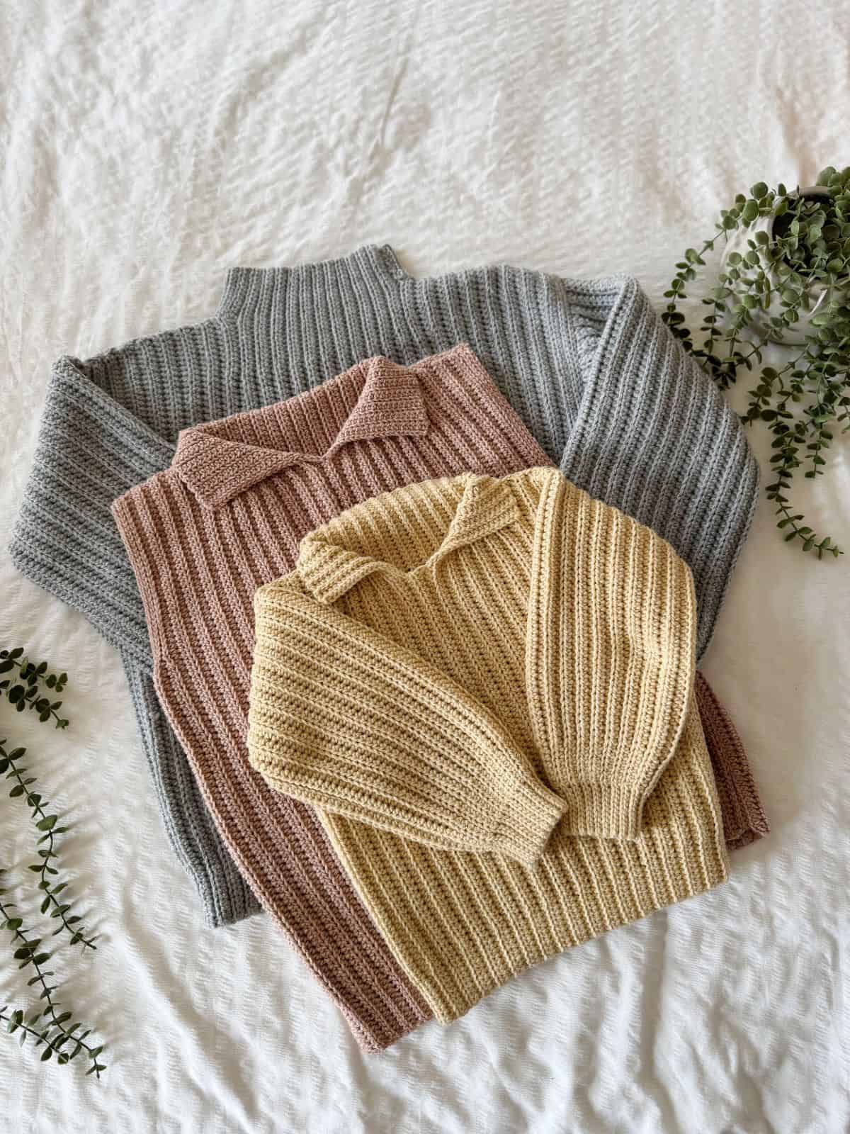Three ribbed crochet sweaters in gray, pink, and yellow are neatly stacked on a white textured surface with small green leafy branches nearby, perfect for pairing with your favorite modern crochet vest pattern.