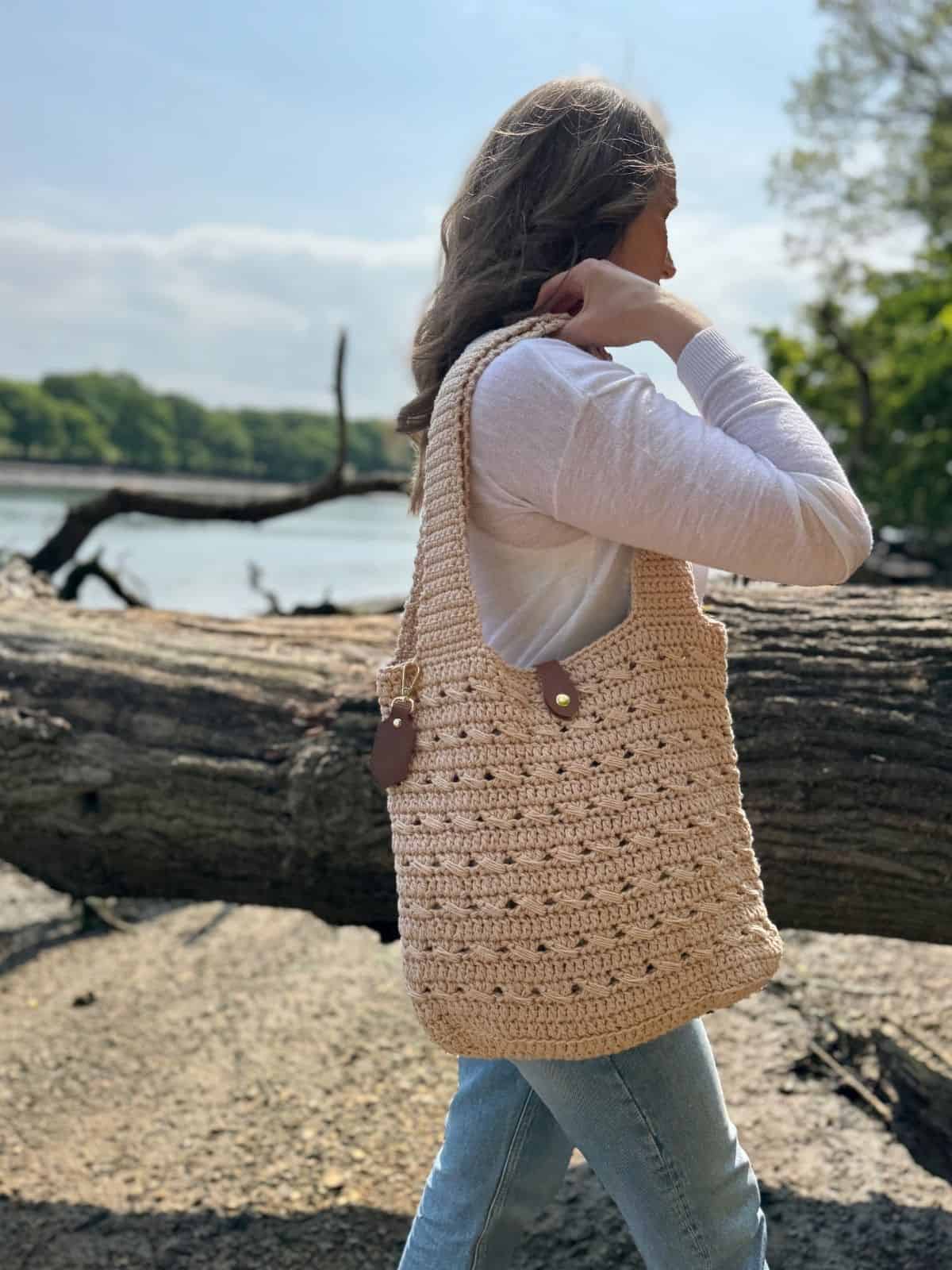 A person wearing a white long-sleeve shirt and jeans stands outdoors by a fallen tree, carrying a large beige tote bag made from a free crochet market bag pattern.