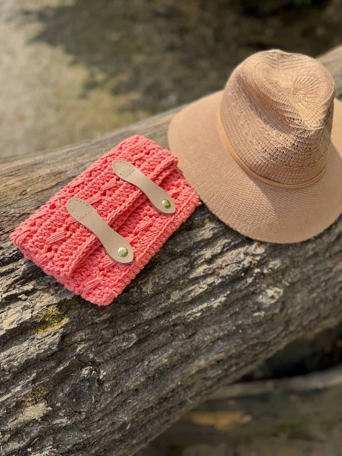 A pink crochet clutch bag pattern with leather straps and a beige sun hat rest on a textured tree trunk outdoors.