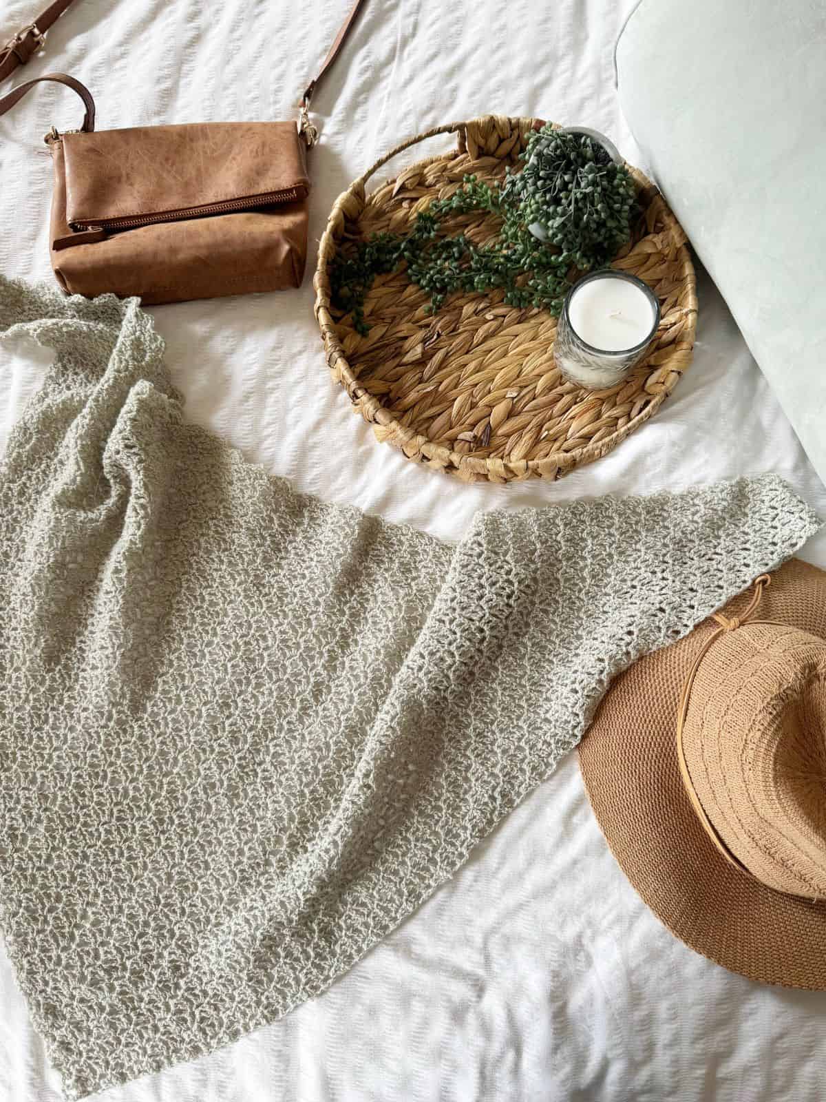 A woven tray with a candle and greenery, a brown bag, a lace crochet shawl pattern in beige, and a straw hat are arranged on a white textured surface.