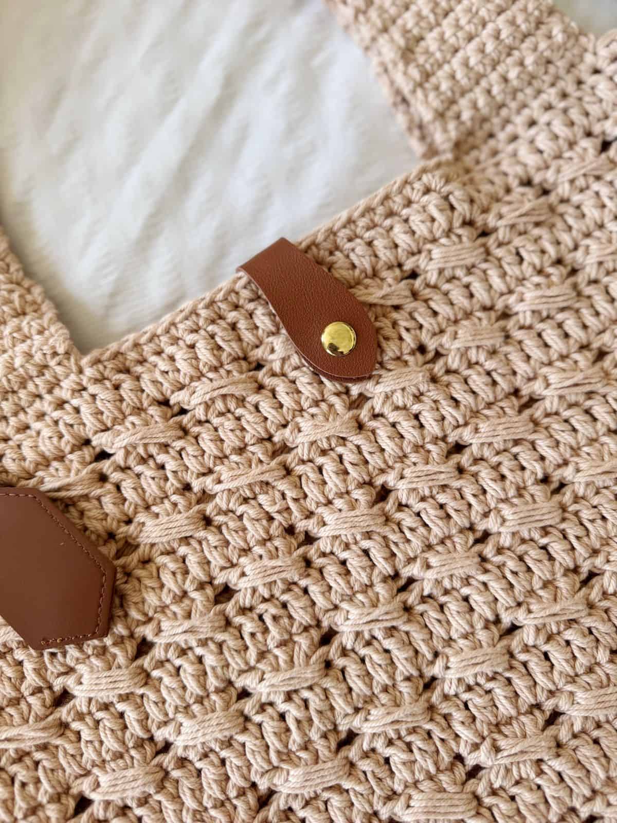 Close-up of a beige crocheted bag with a textured pattern, featuring tan faux leather straps secured by gold-colored buttons. Inspired by a free crochet market bag pattern, it blends style and practicality.