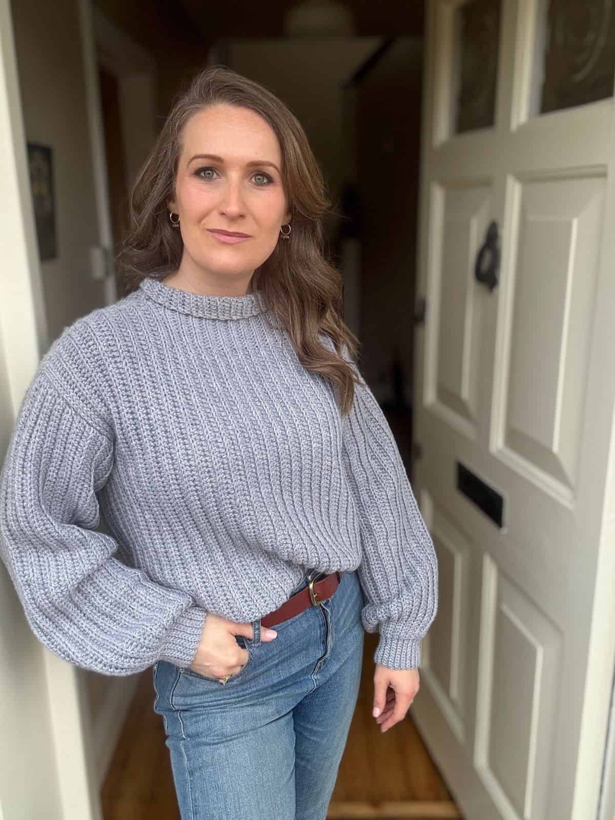 A woman wearing a grey crochet ribbed sweater pattern and blue jeans stands in a doorway with one hand in her pocket, looking at the camera.