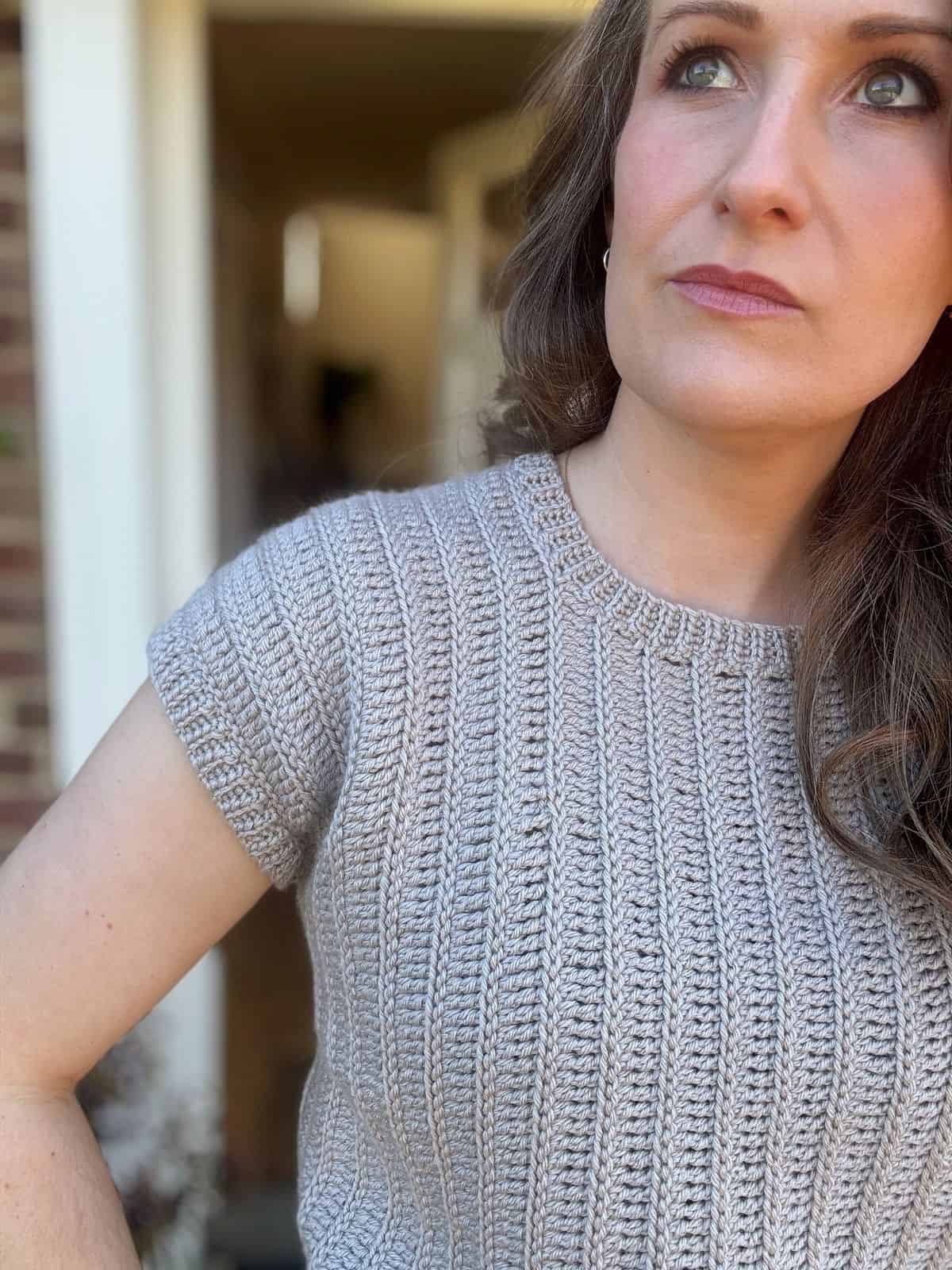 A woman with wavy brown hair wearing a short-sleeve gray crochet summer top stands outdoors, looking slightly upward to her right.