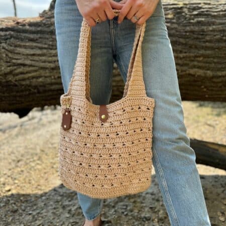 A person wearing blue jeans and brown loafers holds a beige crocheted tote bag with leather handles—crafted from a free crochet market bag pattern—while standing outdoors in front of a large log.
