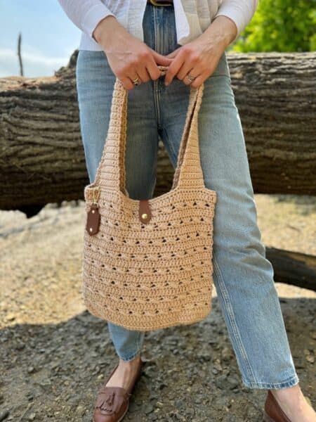 A person wearing blue jeans and brown loafers holds a beige crocheted tote bag with leather handles—crafted from a free crochet market bag pattern—while standing outdoors in front of a large log.