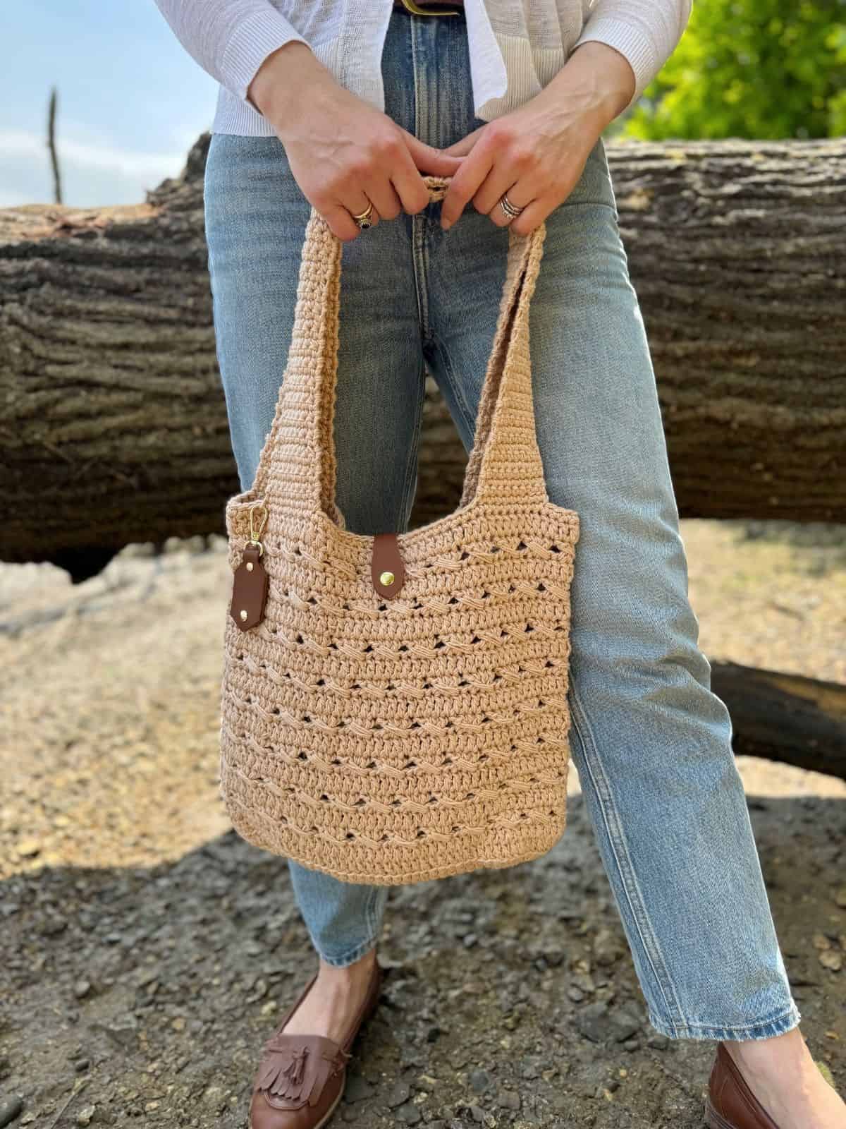 A person wearing blue jeans and brown loafers holds a beige crocheted tote bag with leather handles—crafted from a free crochet market bag pattern—while standing outdoors in front of a large log.