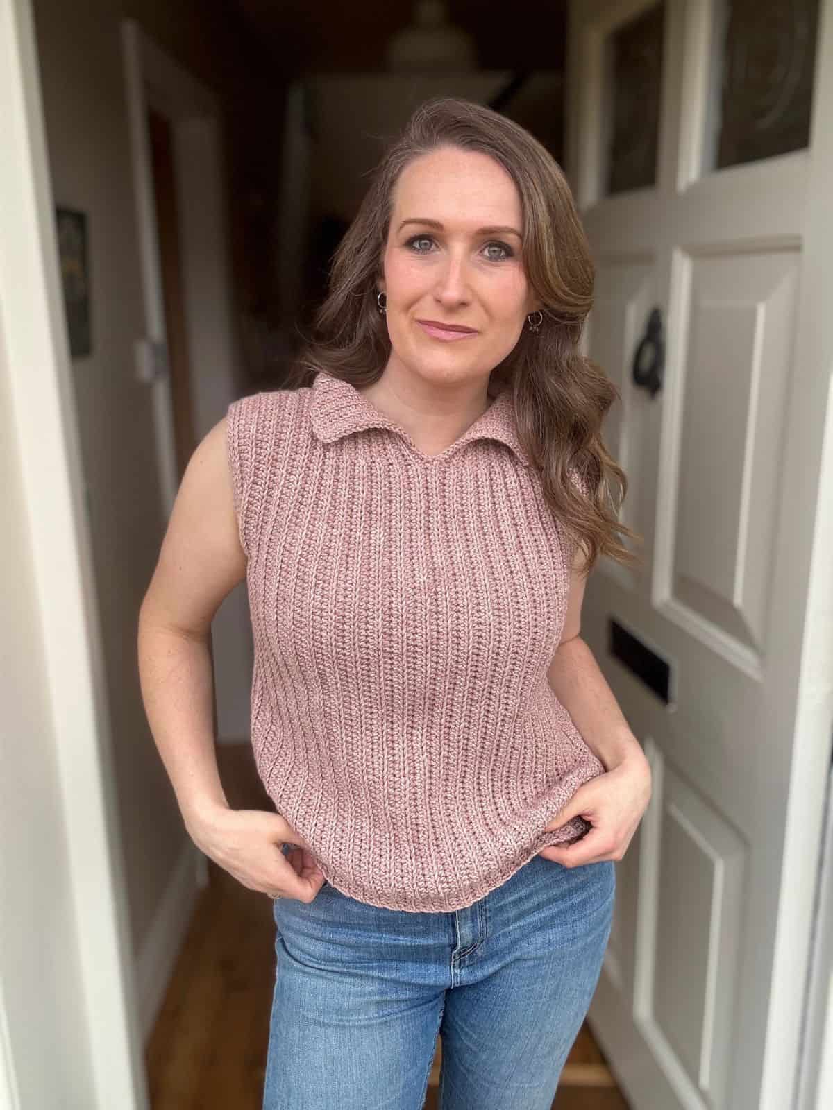 A woman stands indoors in front of a doorway, wearing a sleeveless, textured pink top inspired by a modern crochet vest pattern and blue jeans, looking directly at the camera.