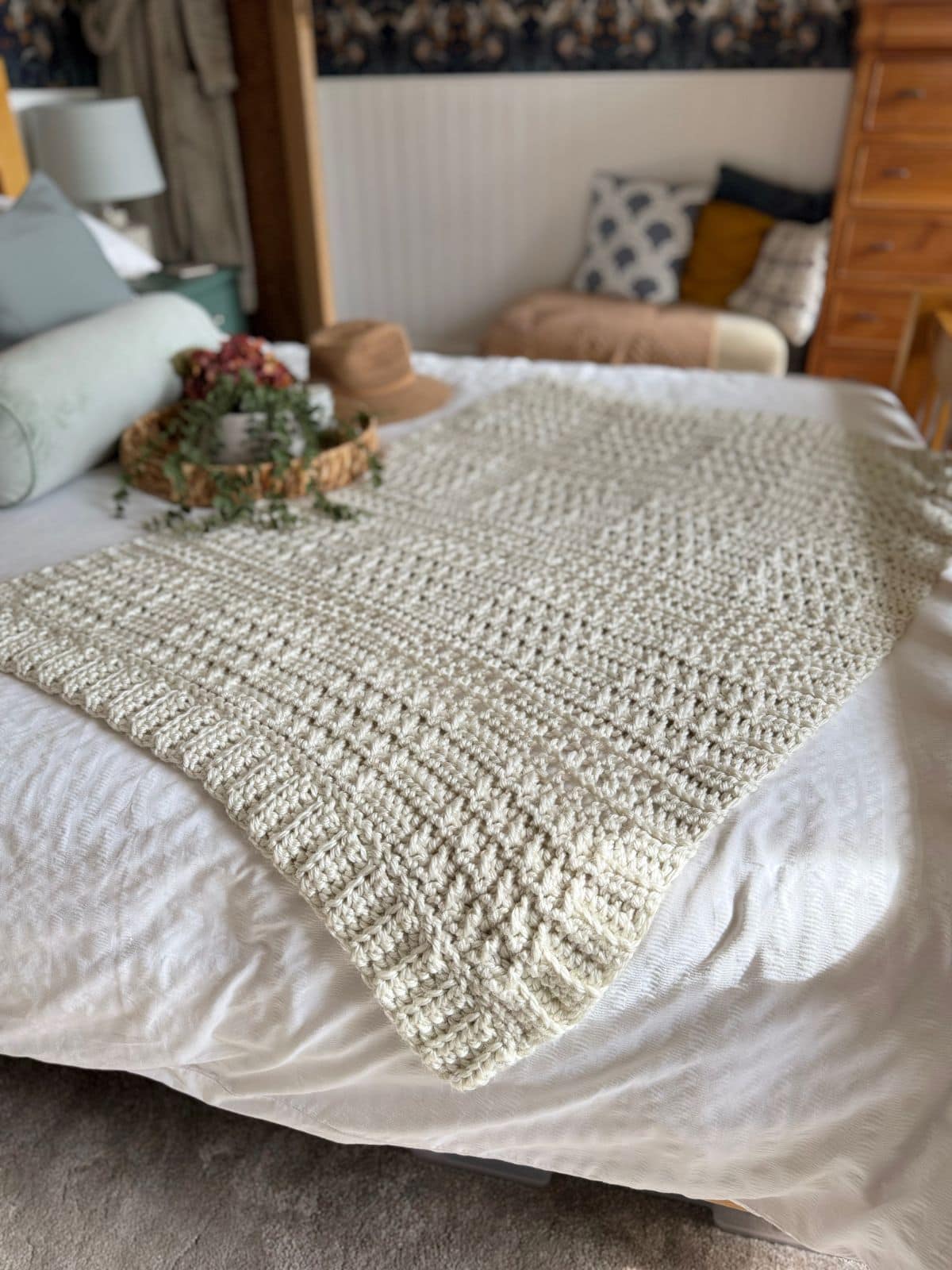 A thick crochet blanket pattern in cream is spread out on a neatly made bed with pillows, a hat, and a woven tray with a plant in the background.