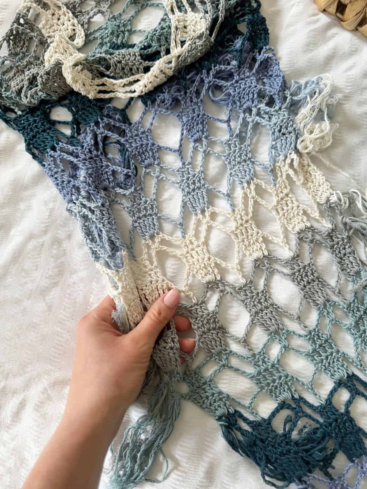 A hand holds a piece of loosely woven, multicolored crochet fabric with blue, green, and cream tones—perfect inspiration for a stylish bandana scarf crochet pattern—set against a white background.