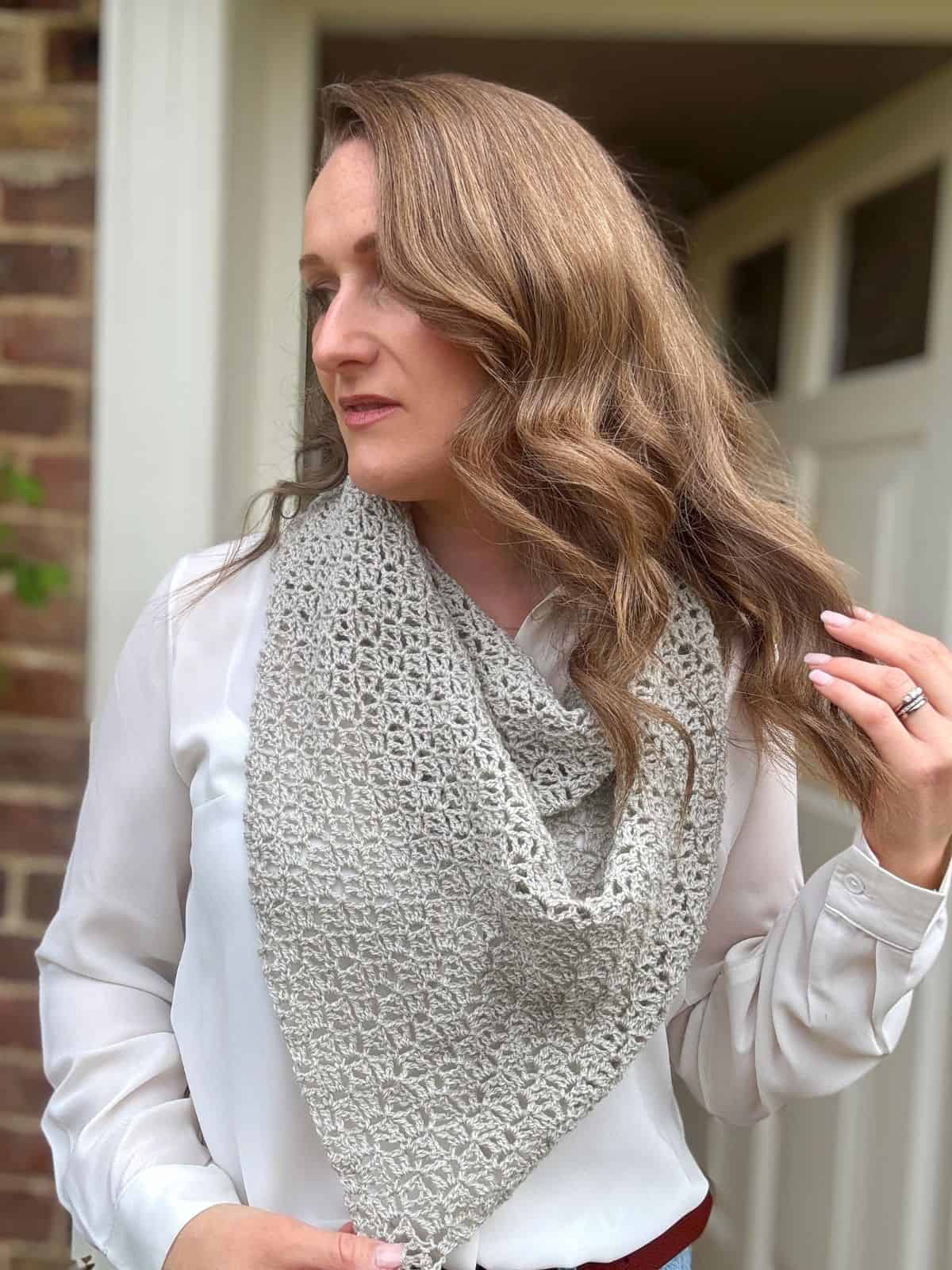 A woman with wavy light brown hair wears a light-colored lace crochet shawl pattern as a scarf over her white blouse, standing in front of a brick wall and white door, looking to her left.