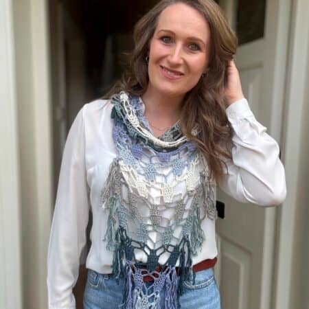 Woman standing in front of an open door, wearing a white blouse, blue jeans, and a multicolored bandana scarf crochet pattern, with one hand touching her hair and smiling at the camera.