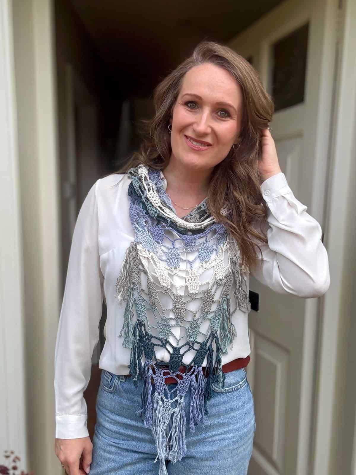 Woman standing in front of an open door, wearing a white blouse, blue jeans, and a multicolored bandana scarf crochet pattern, with one hand touching her hair and smiling at the camera.