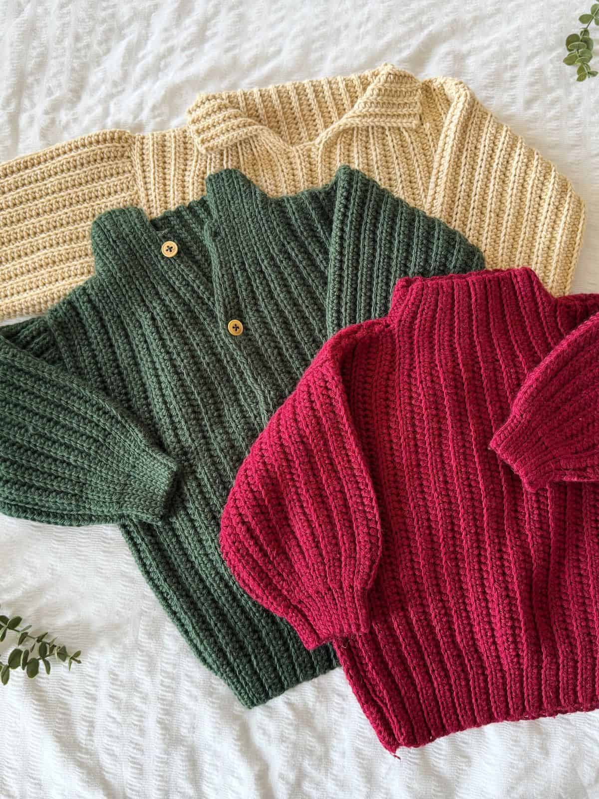 Three ribbed crochet sweaters in cream, green, and red are laid out on a white textured surface. The cozy designs are perfect inspiration if you’re searching for easy crochet sweater patterns for beginners. The green sweater features two collar buttons.