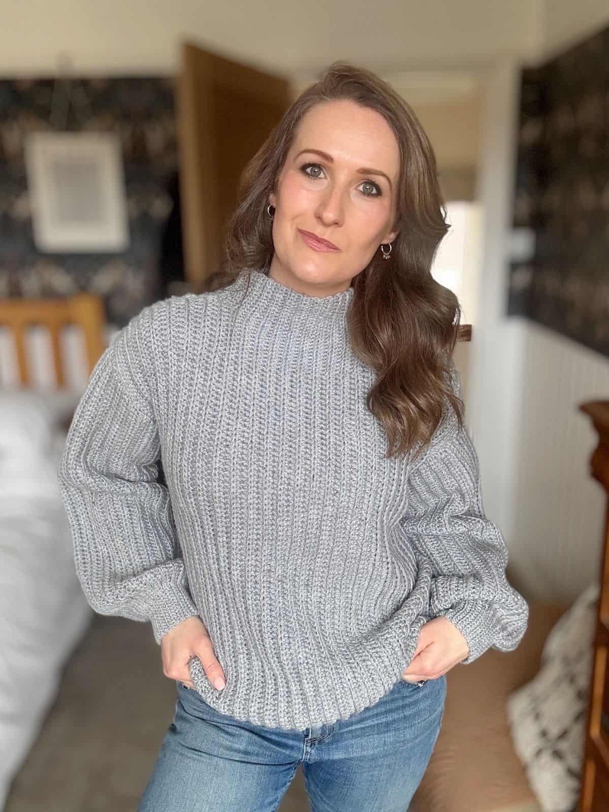 A woman with long brown hair, wearing a gray crochet sweater and blue jeans, stands indoors with her hands in her pockets.