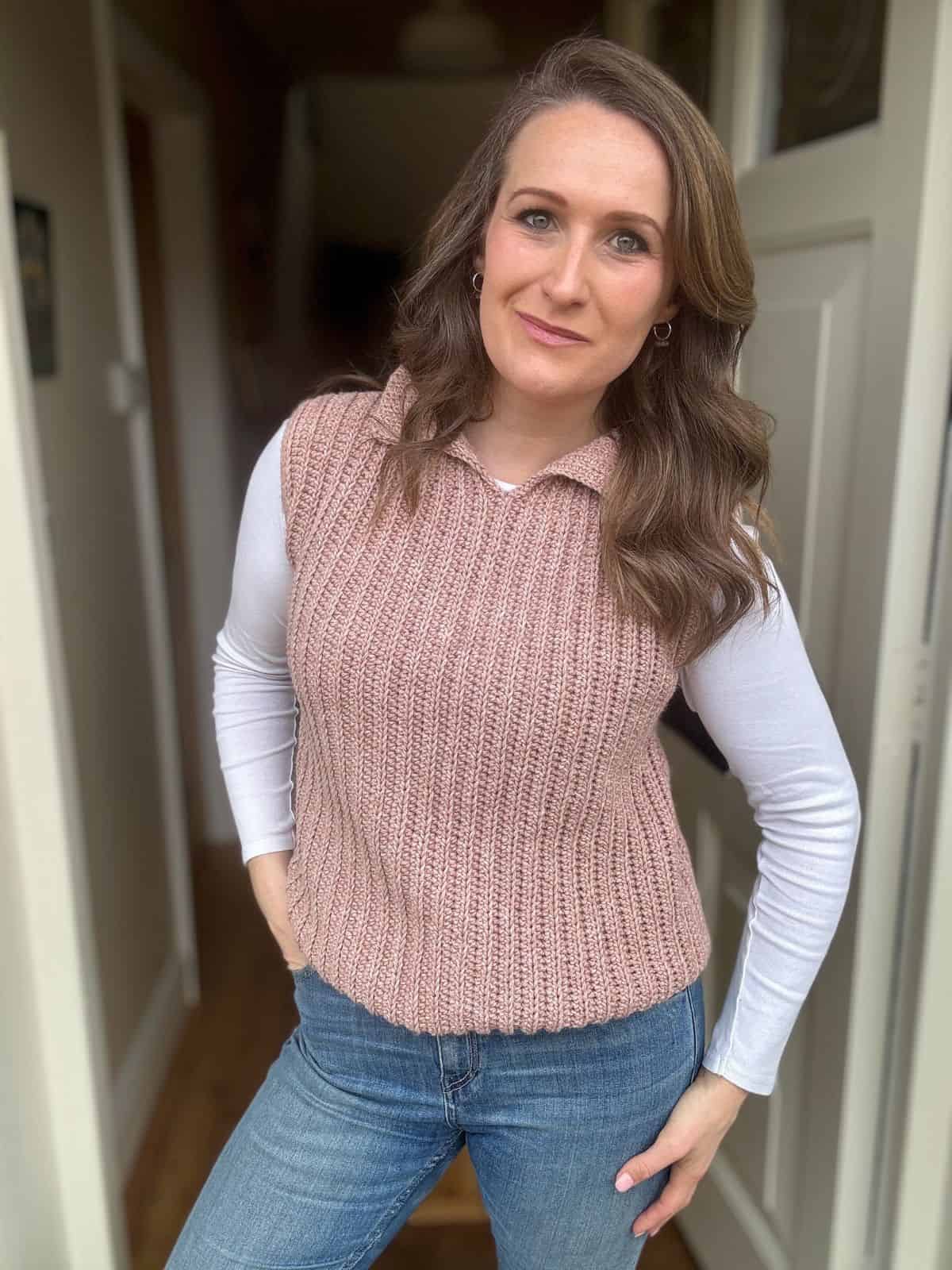 Woman with wavy brown hair wears a light pink modern crochet vest pattern over a white long-sleeve shirt and blue jeans, standing indoors with one hand on her hip.
