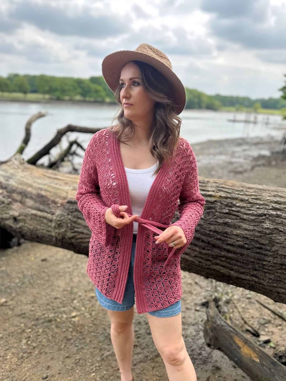 Woman wearing a pink summer cardigan crochet pattern, white top, denim shorts, and a brown hat stands by a large fallen tree near a riverbank on a cloudy day.
