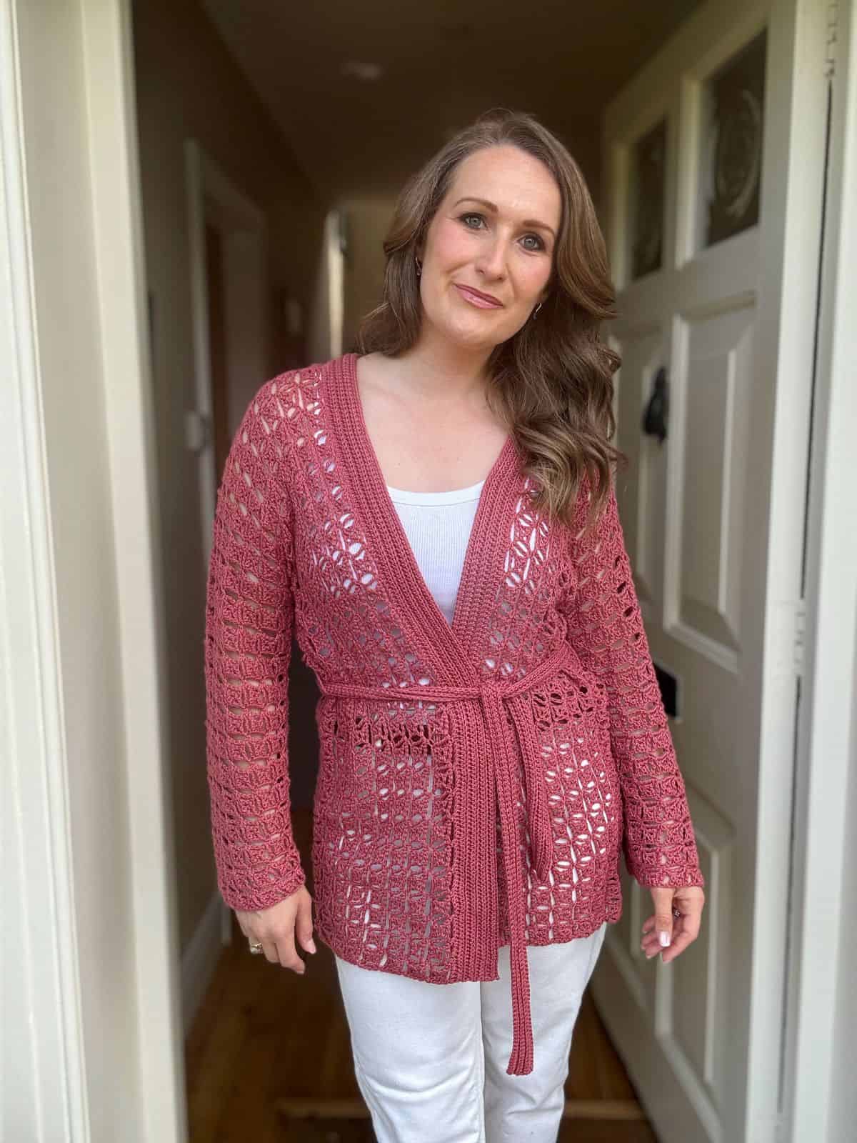 A woman stands in a hallway wearing a pink open crochet cardigan with a belt over a white outfit, looking at the camera with a neutral expression.