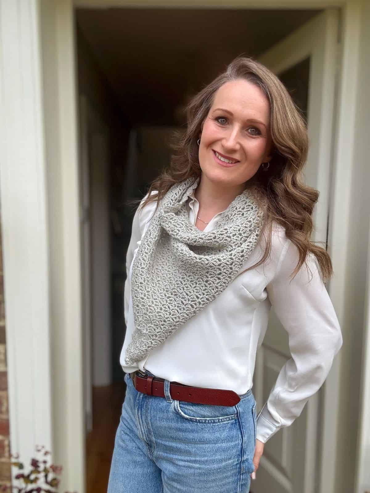 A woman with wavy brown hair, wearing a white blouse, blue jeans, a red belt, and a light gray crochet scarf, stands smiling in a doorway.