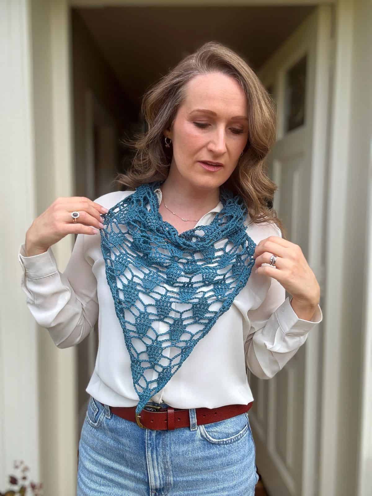 A woman stands indoors wearing a cream blouse and jeans, displaying a blue bandana scarf crochet pattern draped stylishly around her neck.