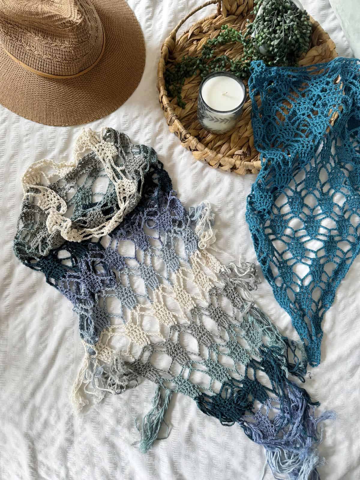 A crocheted scarf and shawl in blue and cream tones, inspired by a bandana scarf crochet pattern, are laid on white fabric next to a straw hat, a woven tray with greenery, and a white candle.